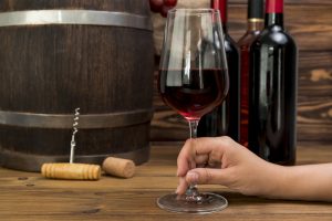A hand holding a glass of red wine beside wine bottles and a barrel