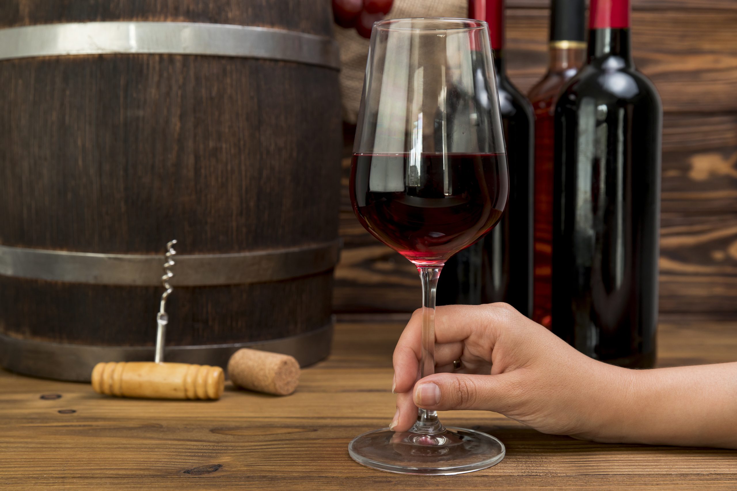 A hand holding a glass of red wine beside wine bottles and a barrel